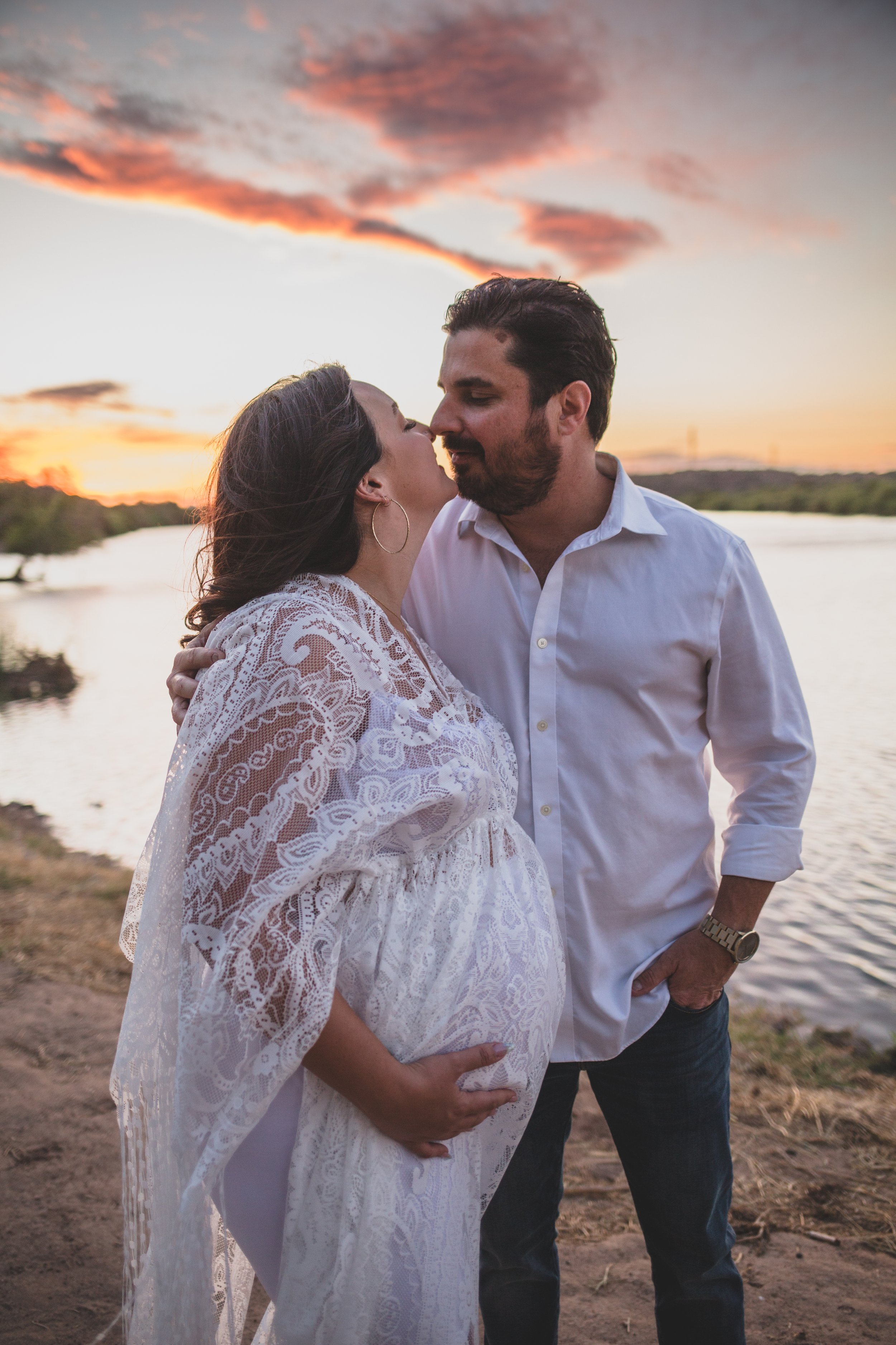 Romantic couple poses with the wind in their hair for maternity photos near the water at sunset the Salt River by timeless Phoenix maternity photographer; Jennifer Lind Schutsky.