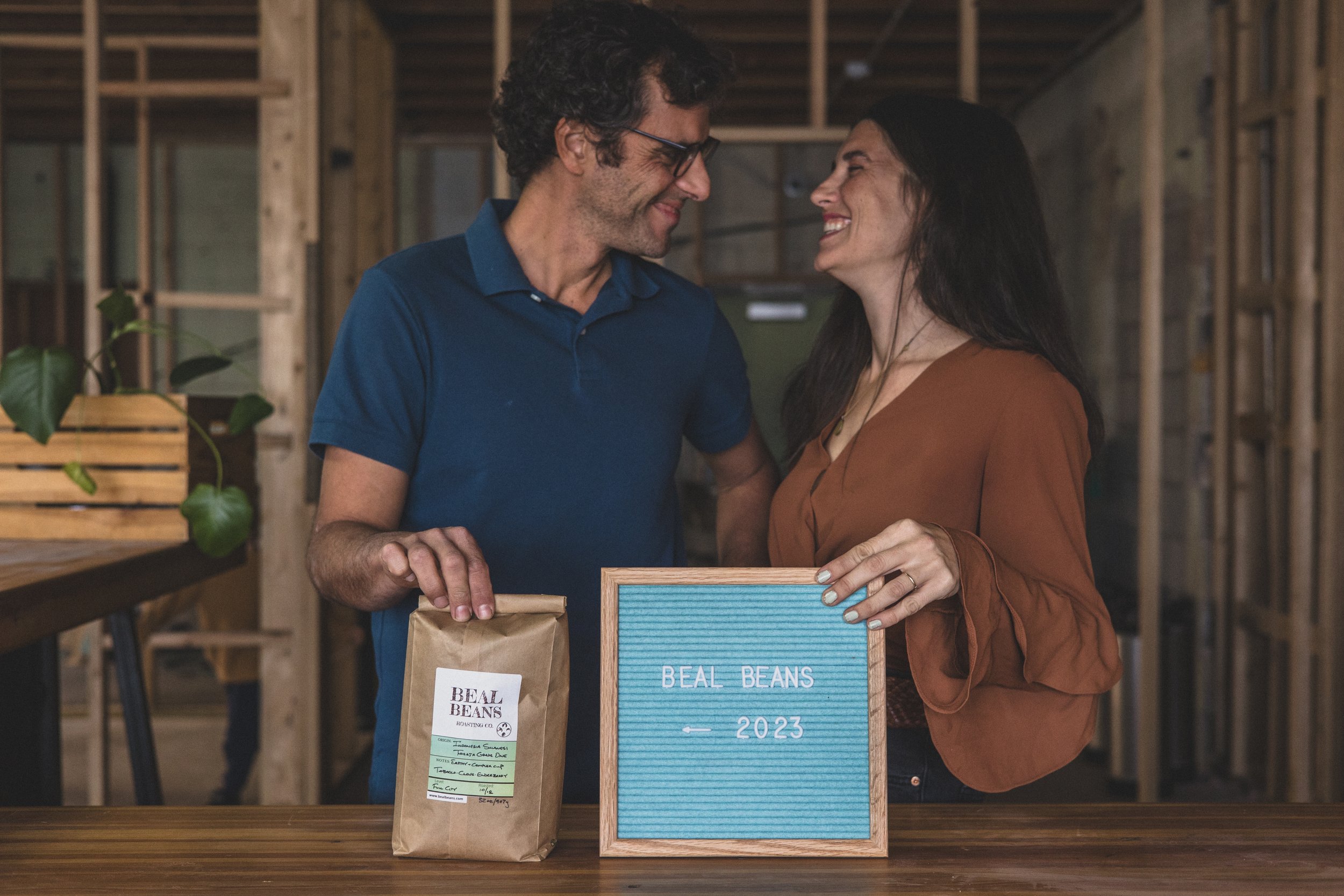 Couple poses with bag of Beal Beans fresh roasted coffee in the unfinished Beal Beans’ new coffee shop coming to Sunnyslope, AZ by Arizona family photographer, Jennifer Lind Schutsky. 