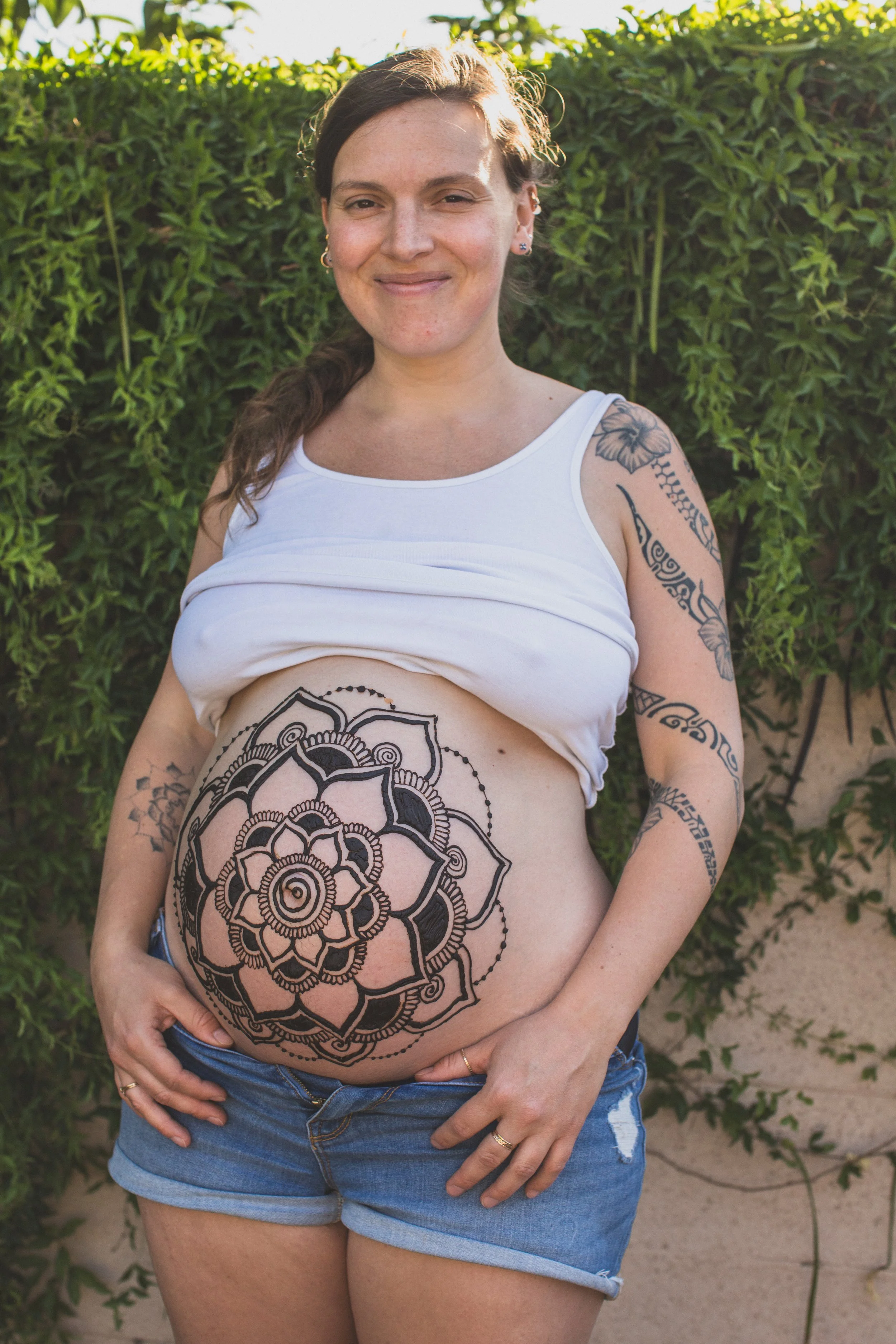 Woman poses in front of plants and outdoor wall in Arizona after receiving maternity henna by traveling henna artist Jennifer Lind Schutsky of La Luna Henna.