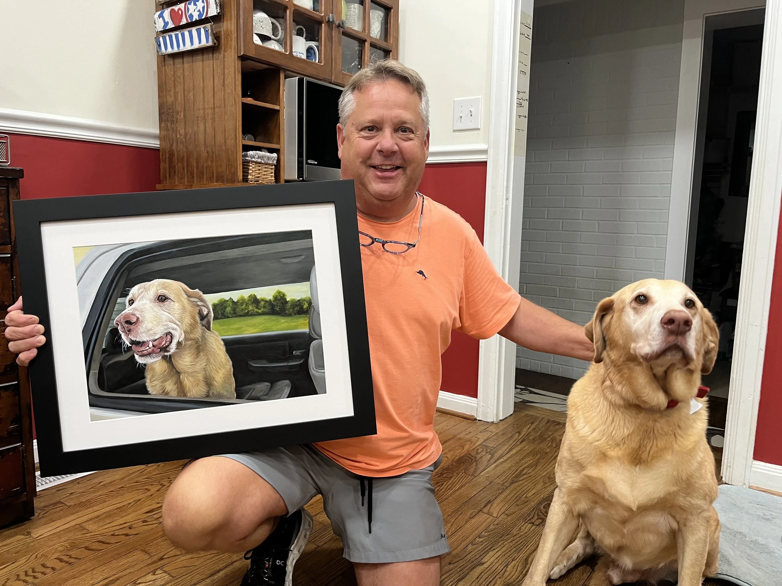A man kneeling on the floor holding a framed portrait of a dog, and sitting next to a real dog.