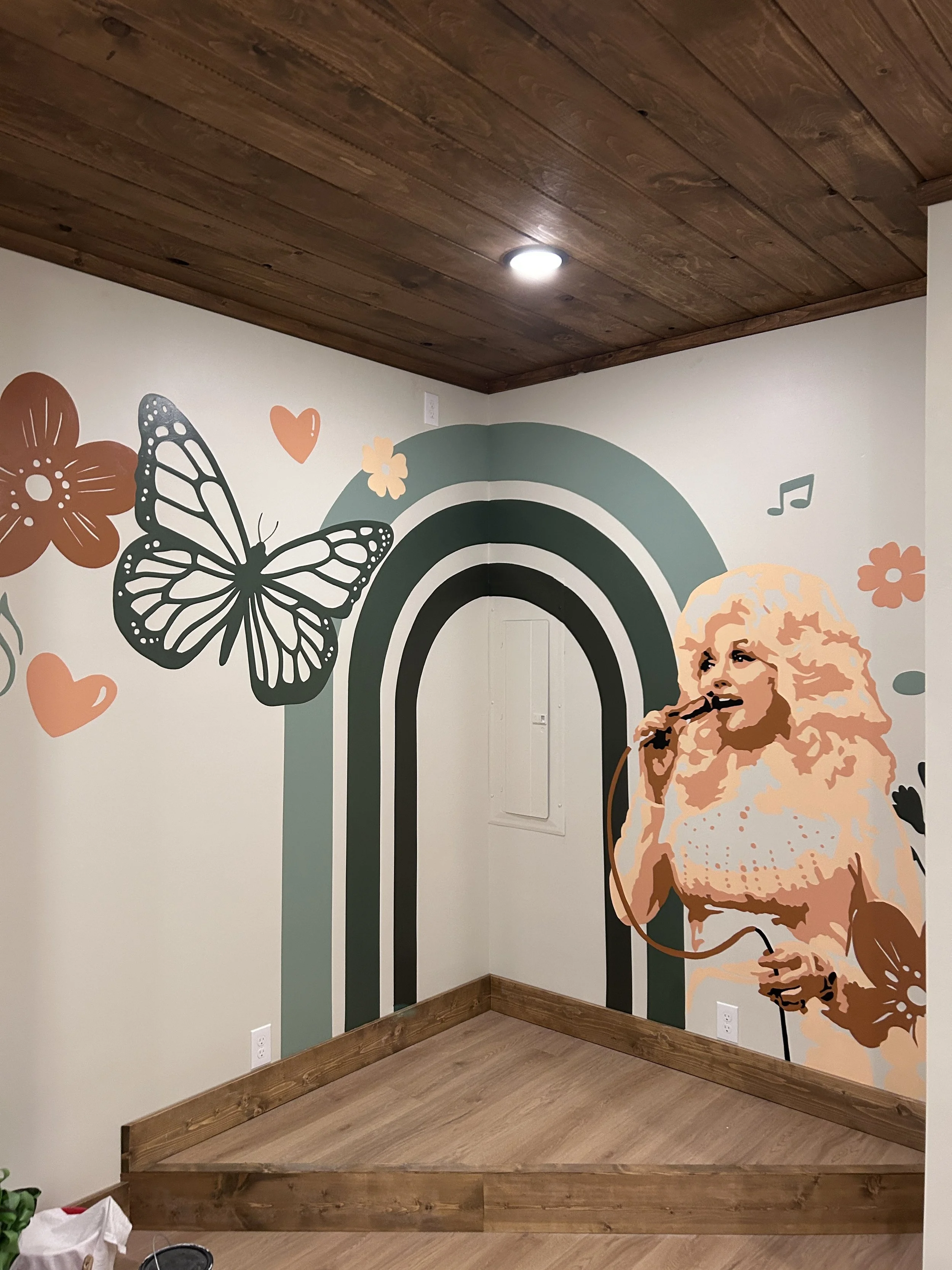 Wall mural with Dolly Parton singing into a microphone, surrounded by butterflies, flowers, hearts, and musical notes, with a rainbow and wooden ceiling. Great for Airbnb walls