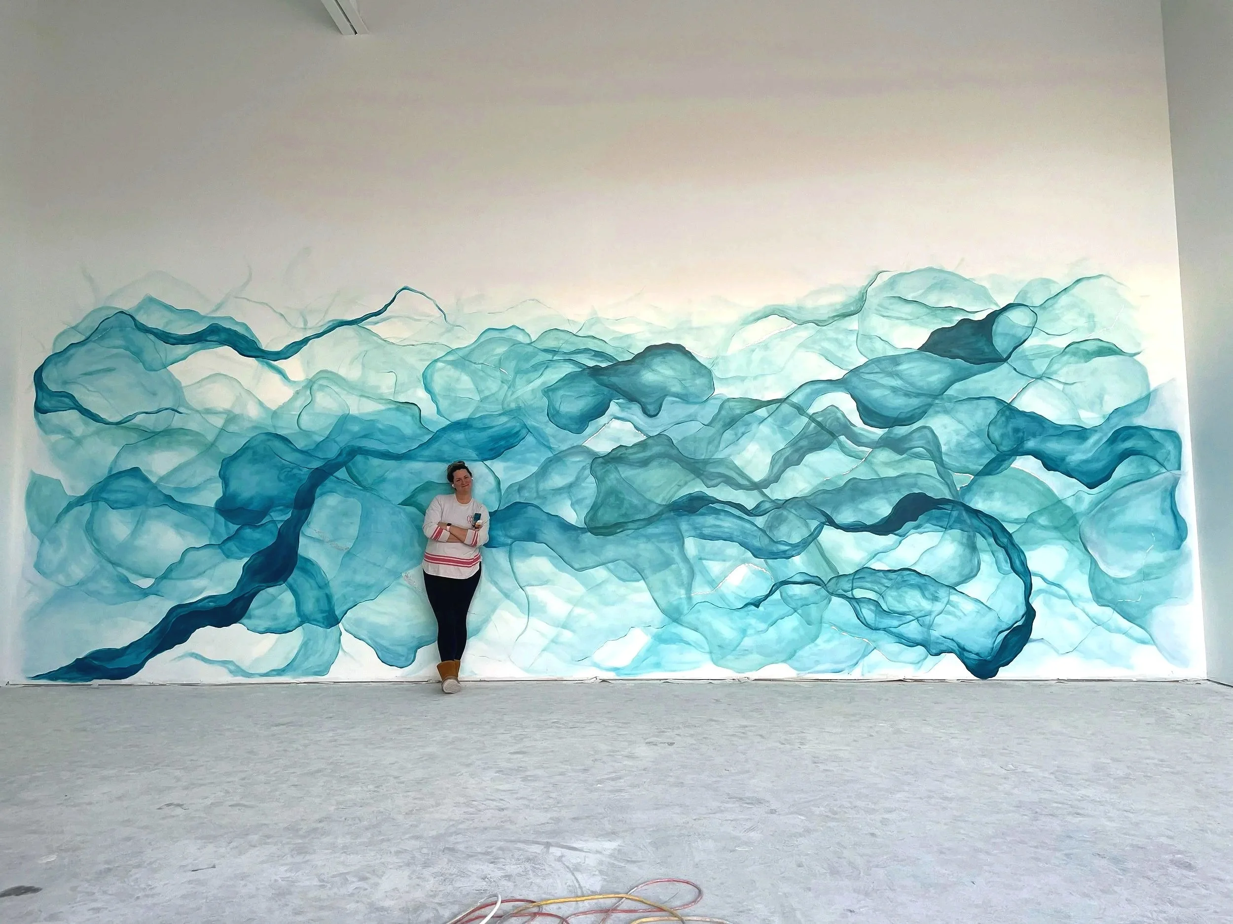 A woman standing in front of a large abstract mural of swirling blue and teal watercolor shapes on a white wall.
