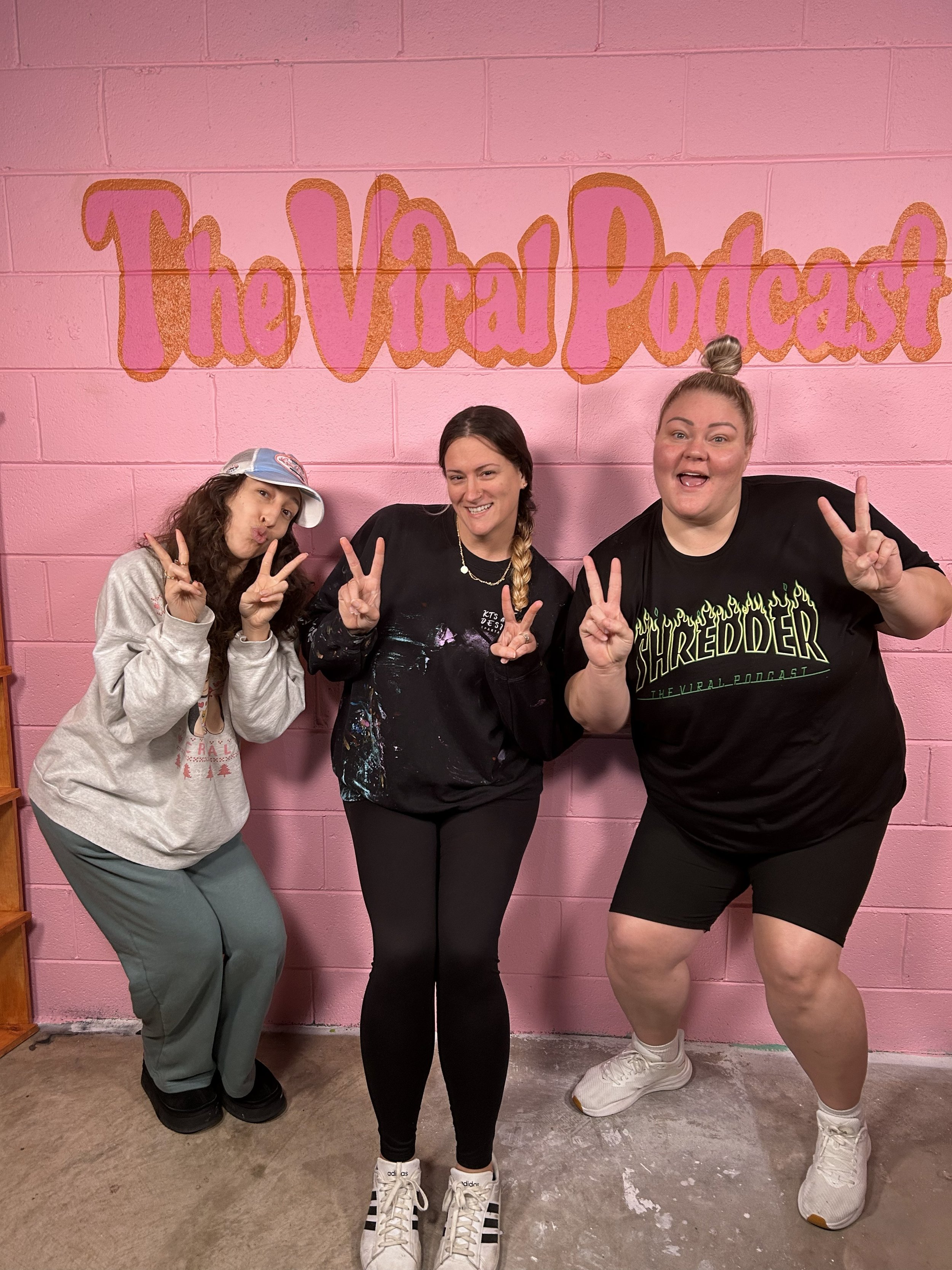 Three women posing with peace signs in front of a pink brick wall with a mural that says 'The Viral Podcast'