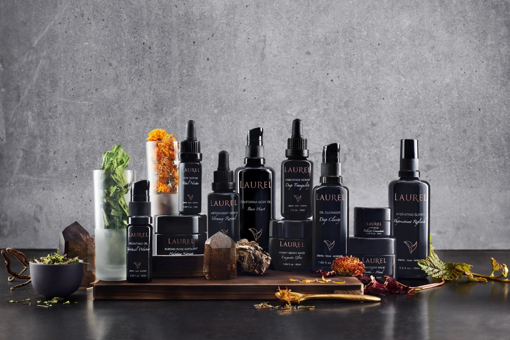 A collection of Laurel skincare products in black bottles and jars with pink and white labels, arranged on a wooden surface with natural elements and decorative rocks, against a gray textured background.