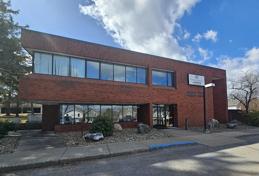 Modern brick building with large glass windows, sign reading 'Sullivan County Medical Center' and 'Laporte', landscaped with rocks and bushes, located in a suburban area under a partly cloudy sky.