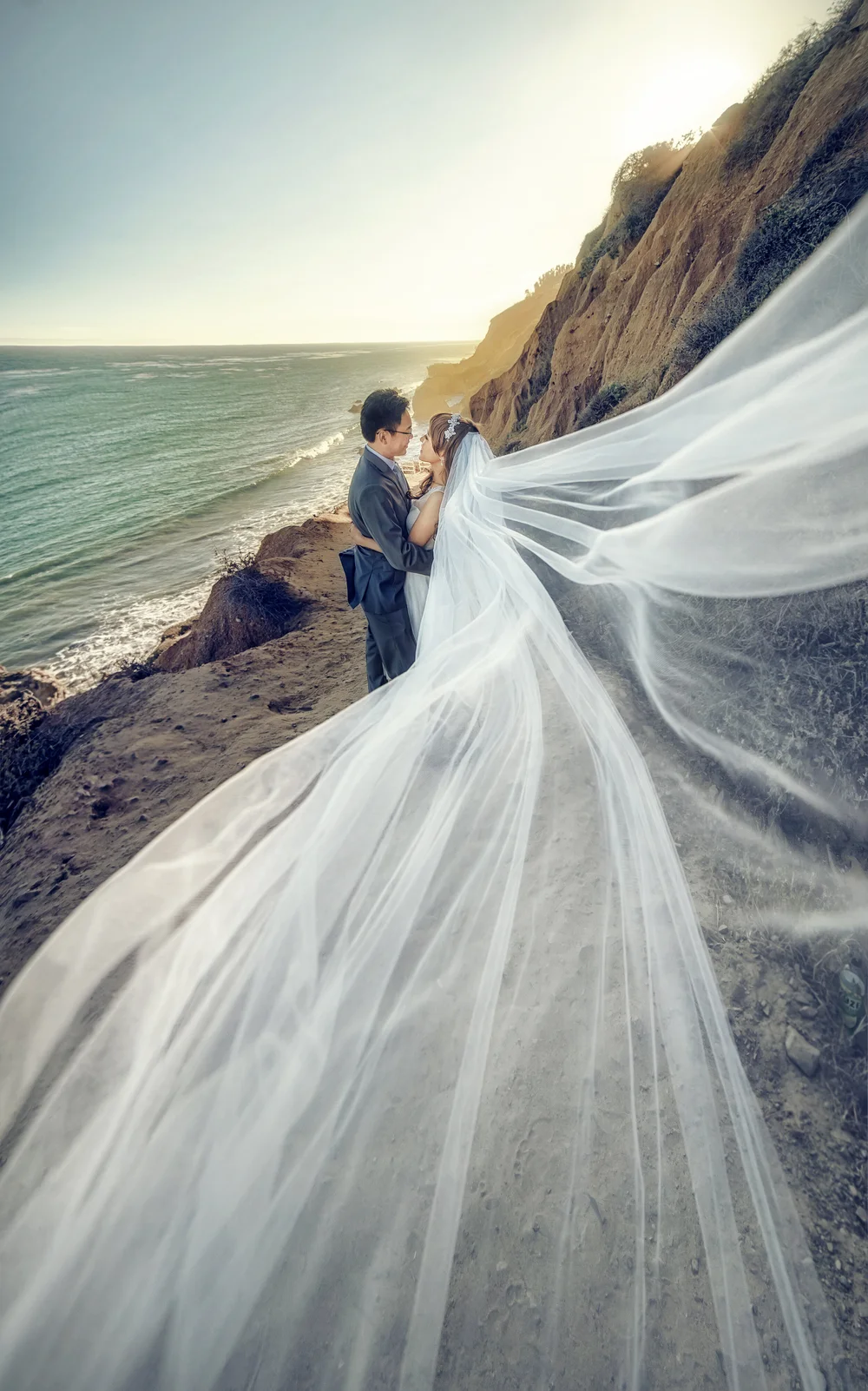 Bride and groom on a coastal bluff, her veil sweeping across the foreground