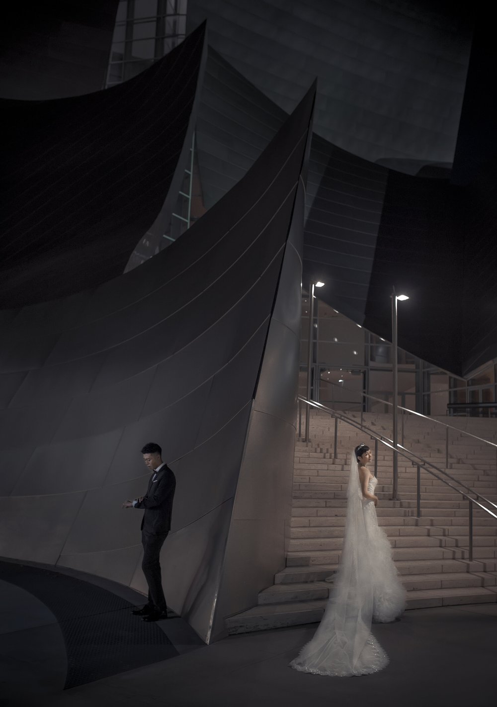 Bride and groom apart on the steps beside the steel curves of a concert hall, black and white