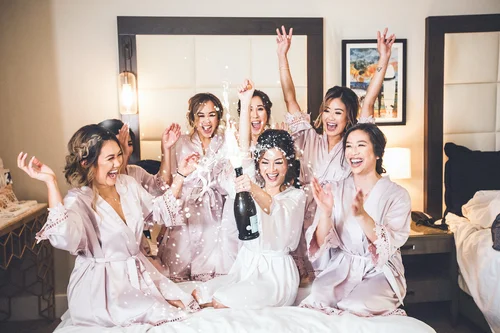 Bride and her bridesmaids in blush robes celebrating with champagne on a hotel bed