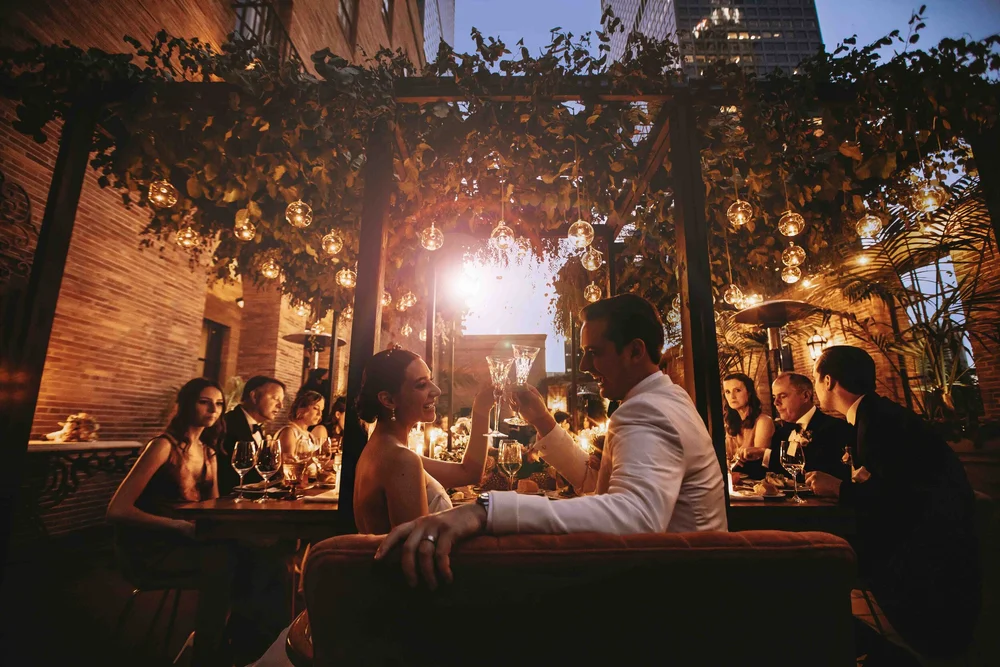Bride and groom on a sofa in a candlelit garden restaurant, guests dining around them