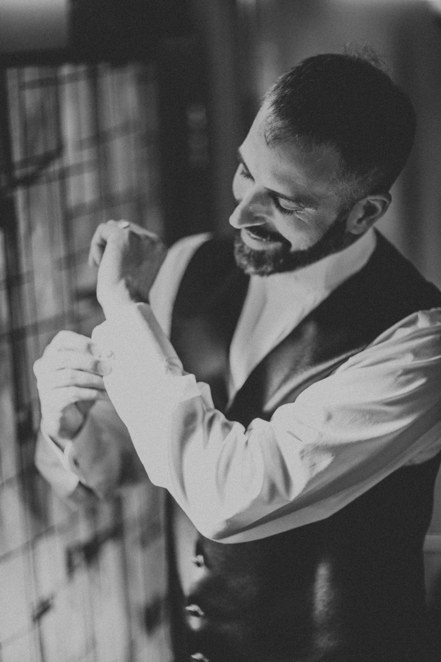 Black and white frame of a groom tying his bow tie beside a window, wearing a waistcoat