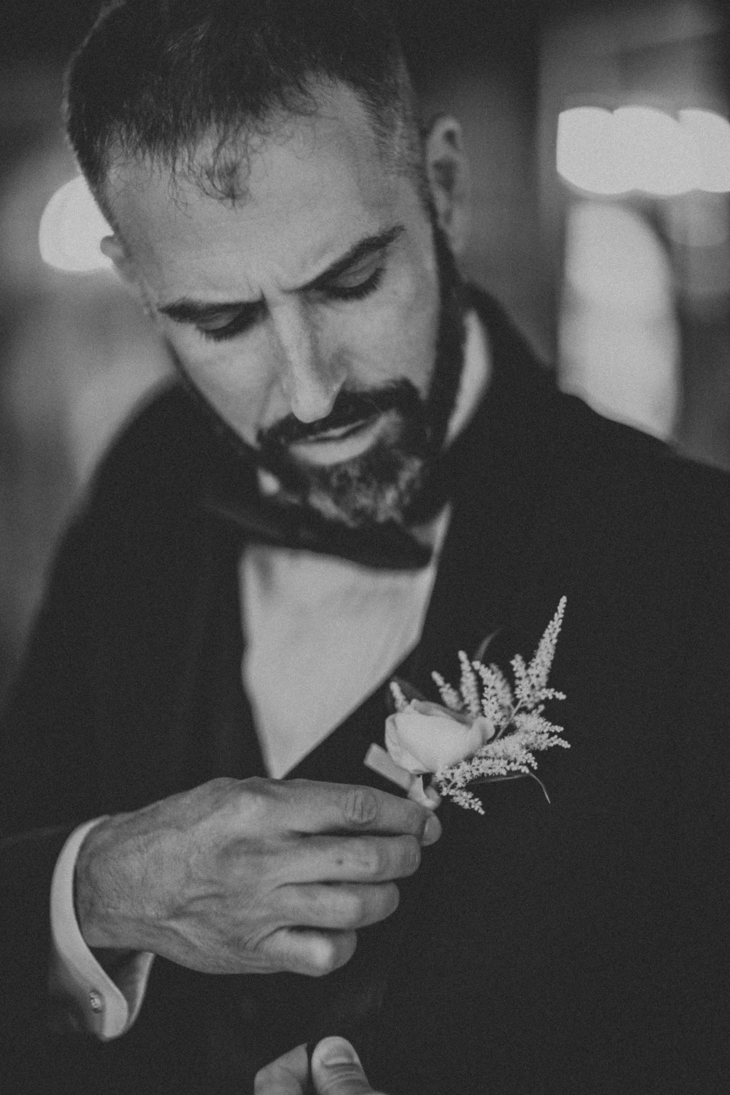 Black and white frame of a groom pinning a white boutonniere to his left lapel