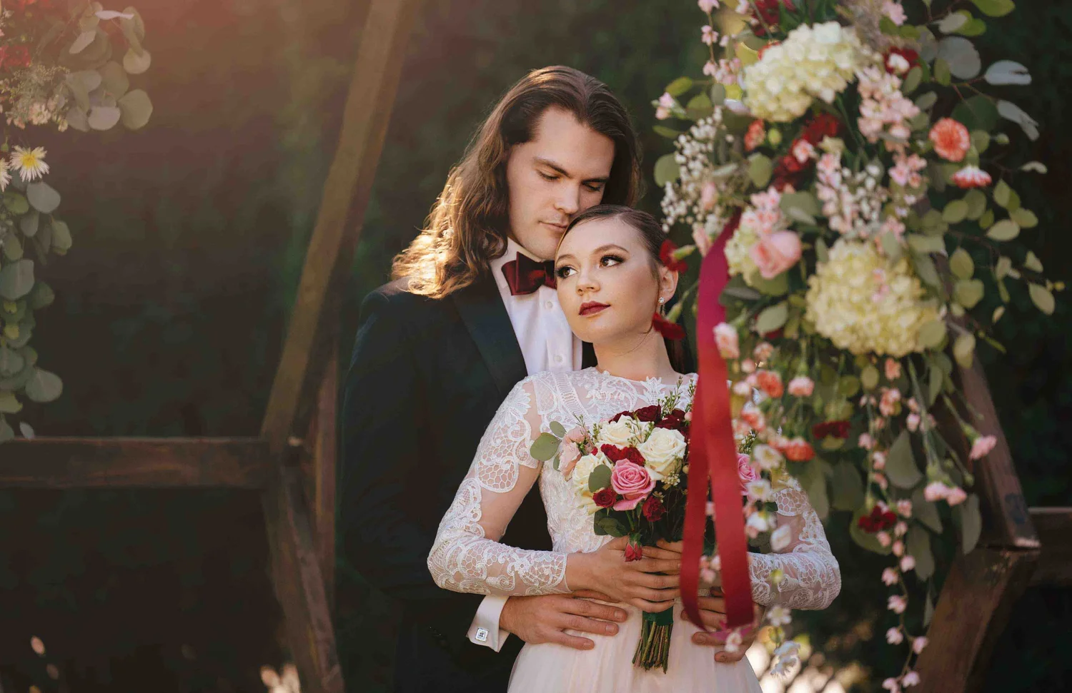 Bride and groom beside a floral arch hung with long red ribbons