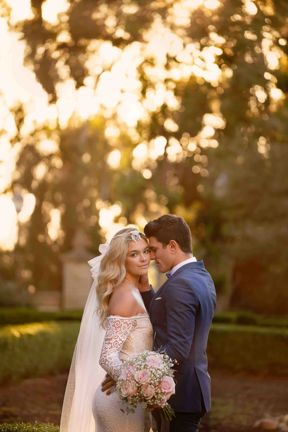 Bride and groom close together under backlit trees, her bouquet between them