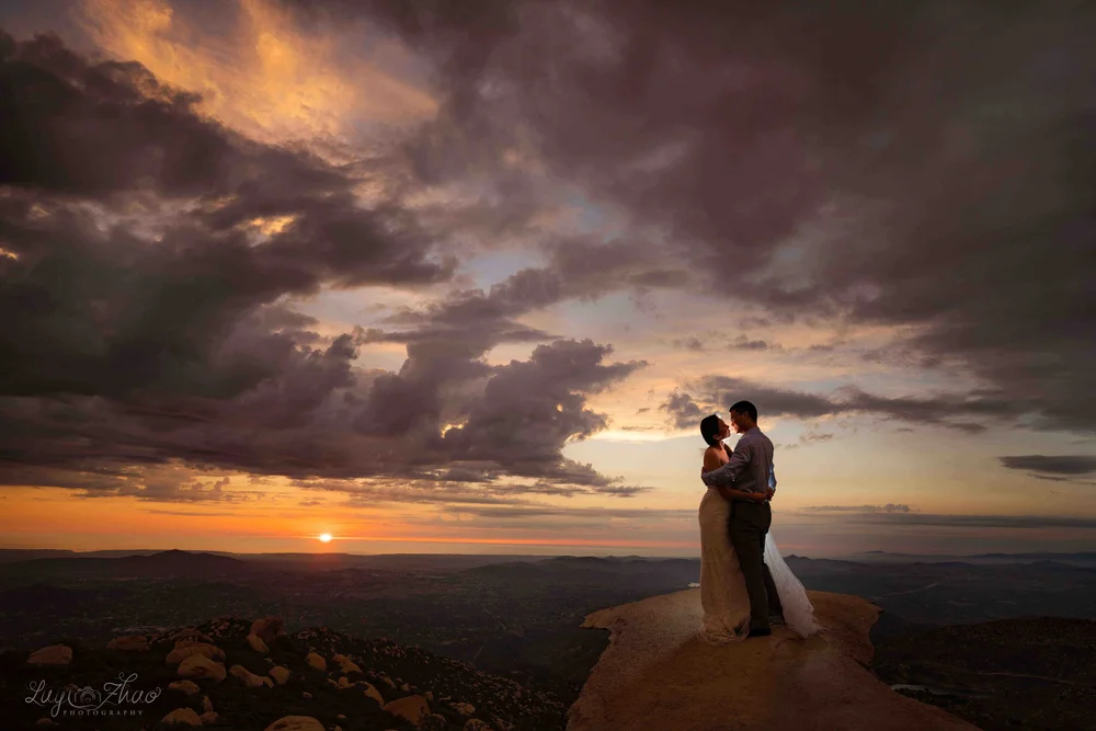A couple in wedding attire standing on a large rock at sunset, embracing and about to kiss with a dramatic sky of clouds and the sun setting over a mountainous landscape in the background.