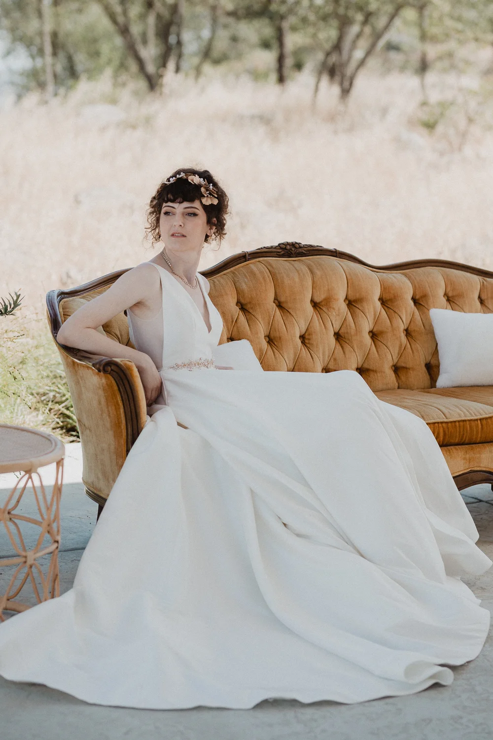 Bride in a flowing gown seated on a vintage velvet sofa against the golden hillside at Oakridge Farmhouse