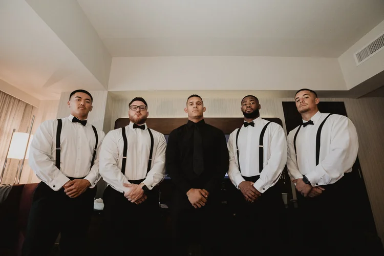 The groom and his groomsmen lined up in white shirts and suspenders in a hotel suite before Anna and Kyle's wedding at City View Penthouse