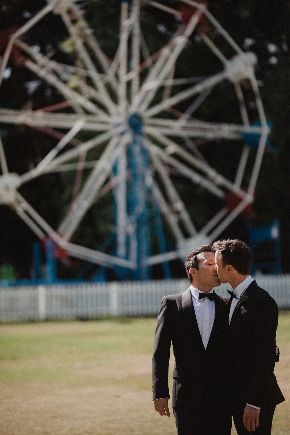 Two grooms in black tie laughing together on a lawn beside a fairground ride