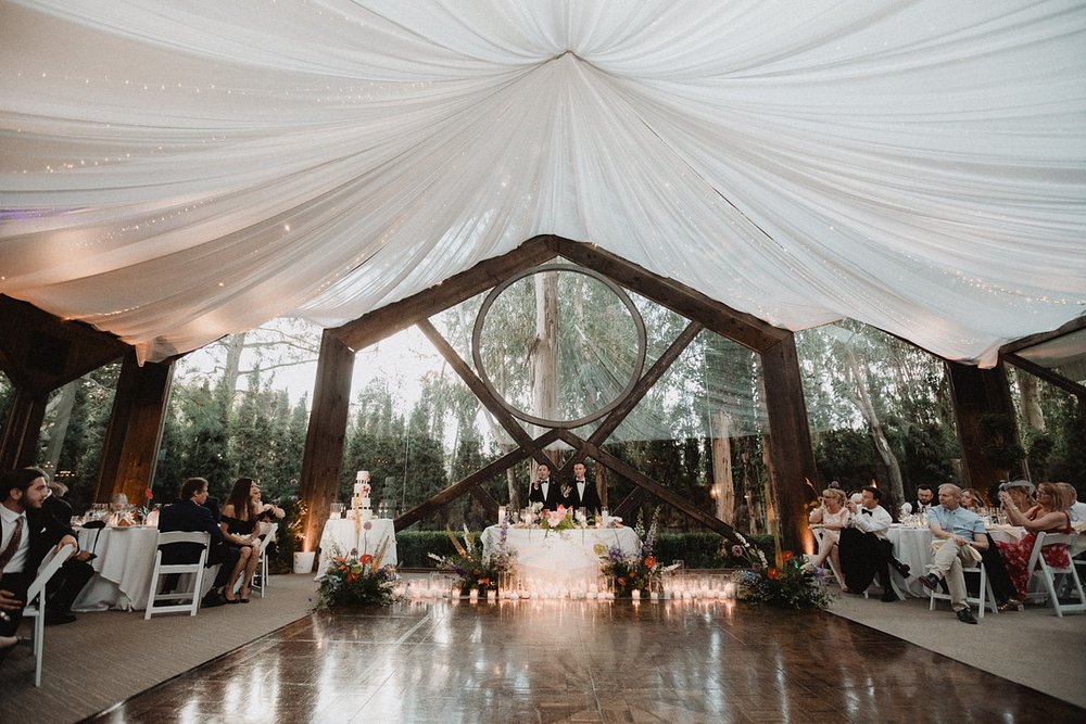 Reception under a draped white tent with a timber-arch frame, tables set around a glowing dance floor