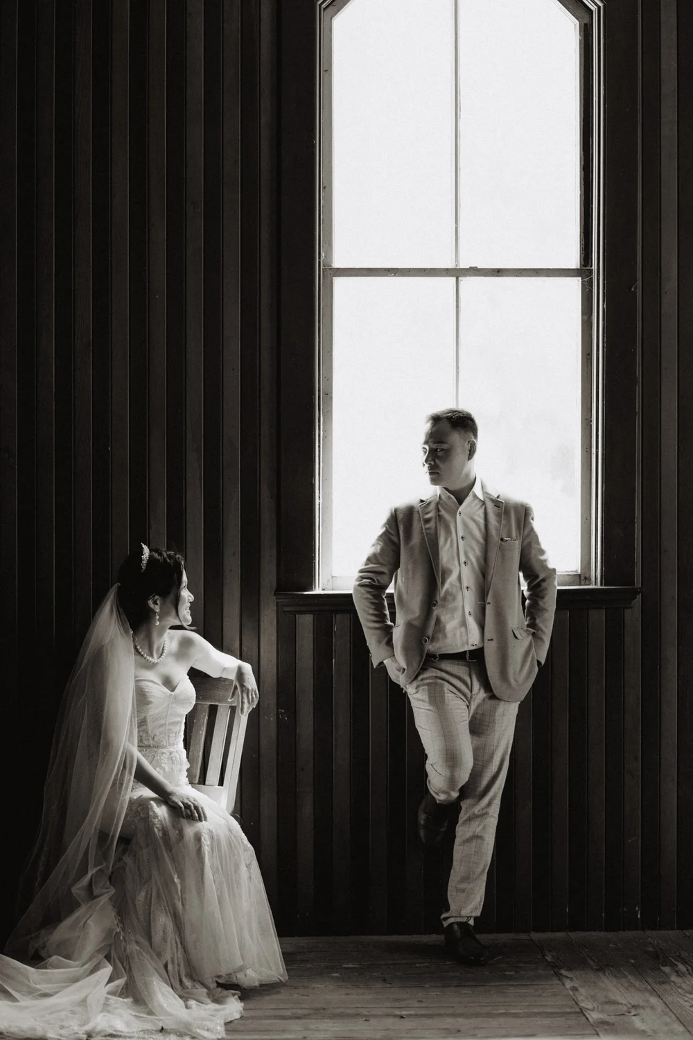 Bride seated and groom standing by a tall window in a dark panelled room, black and white