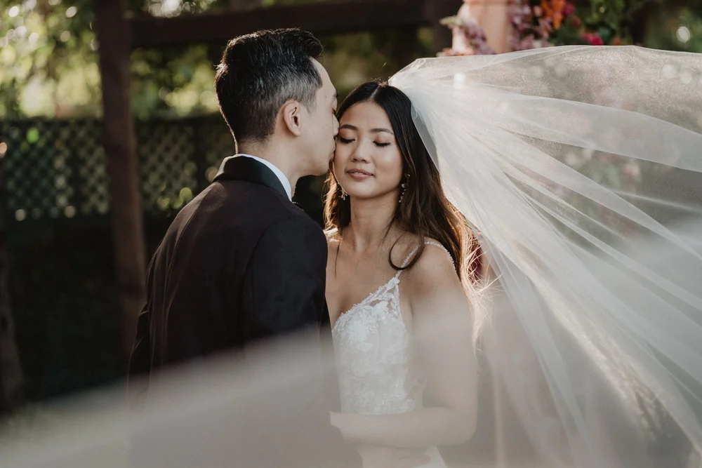 A bride and groom in wedding attire sharing an intimate moment outdoors, with the groom kissing the bride's forehead as her eyes are closed. The bride wears a lace wedding dress and a flowing veil, set against a background of greenery and soft sunlight.