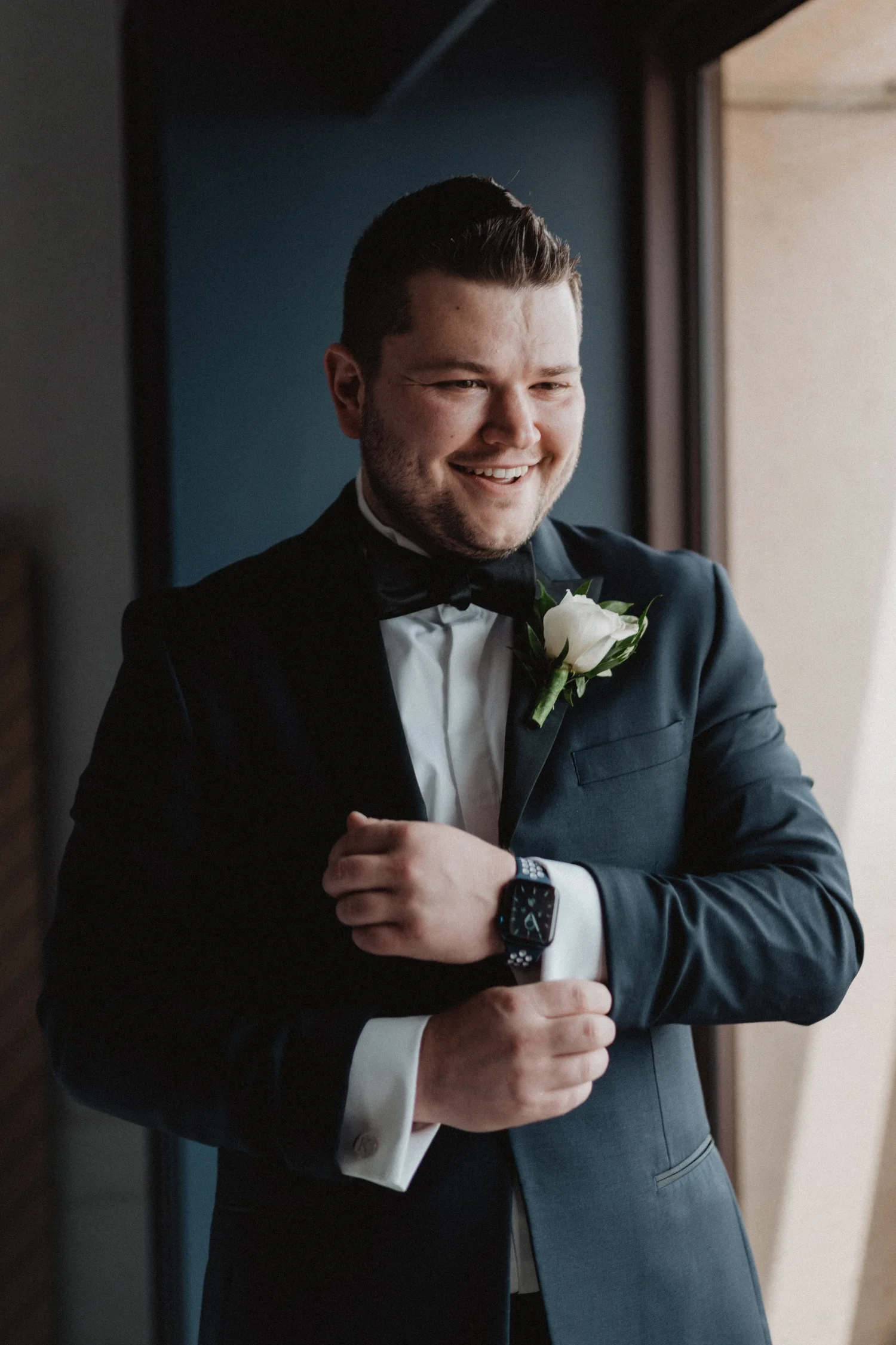 Groom smiling as he fastens his cufflink in a doorway, a white rose boutonniere on his lapel