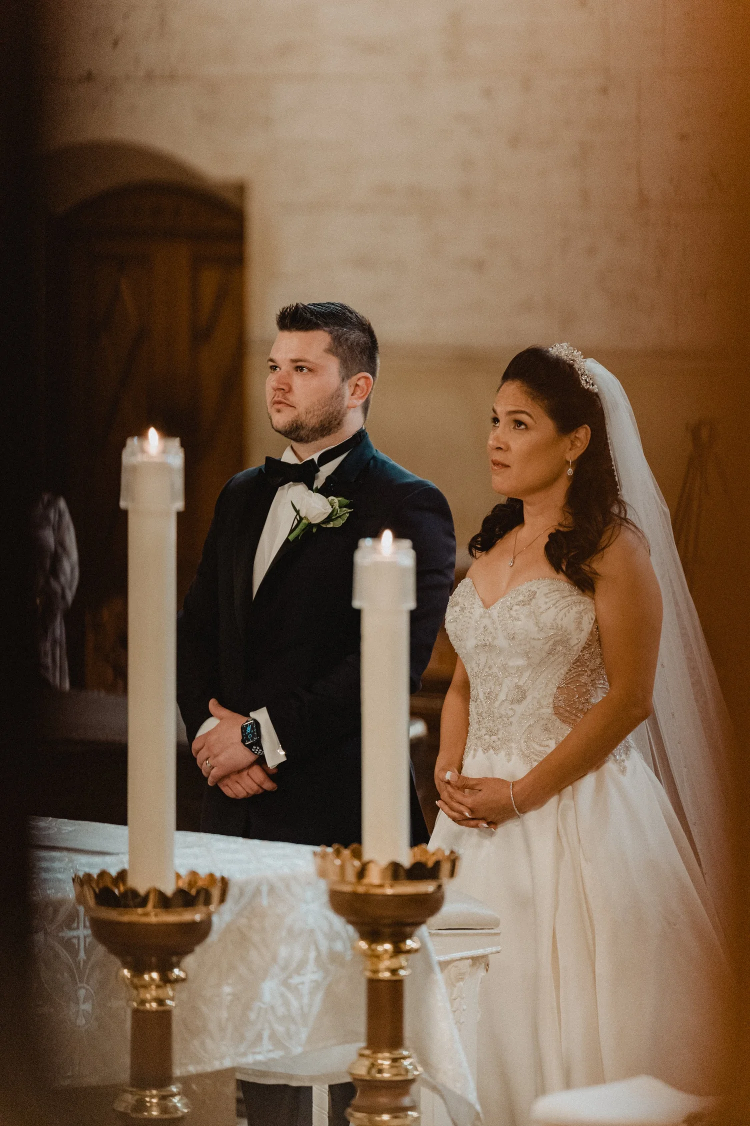 Couple standing at a candlelit church altar, tall pillar candles in gold holders in the foreground