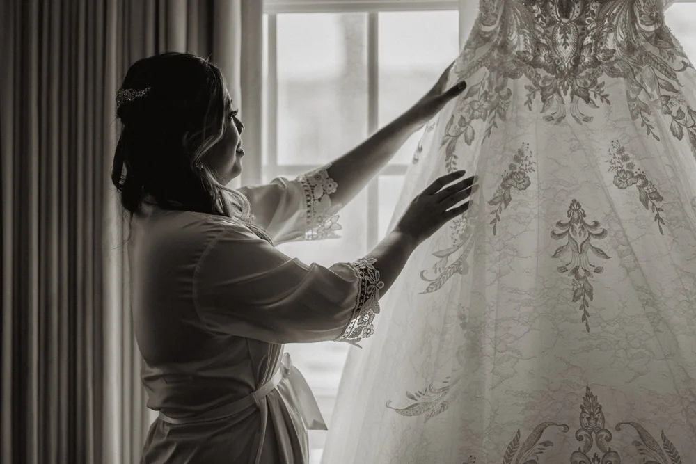 Bride in a robe reaching toward her beaded gown hanging in a window, black and white