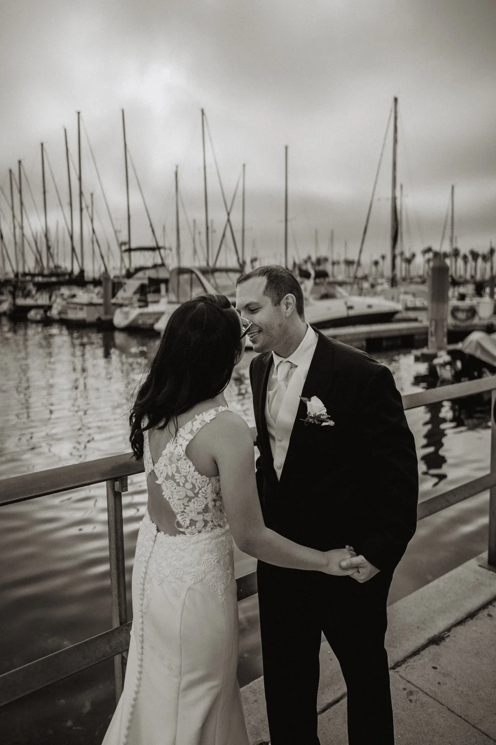 Bride and groom holding hands on a marina dock with masts behind them, black and white