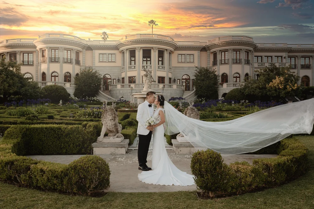 Bride and groom in the formal gardens of a grand estate at sunset, her long veil lifting across the hedges
