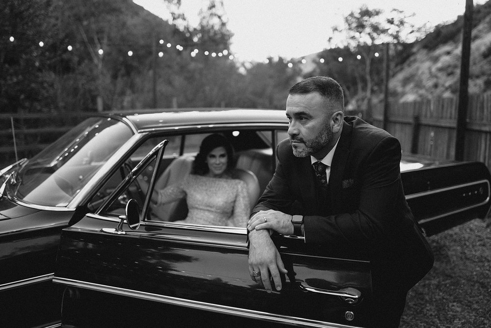 Groom leaning on a vintage car with the bride inside, string lights above, black and white
