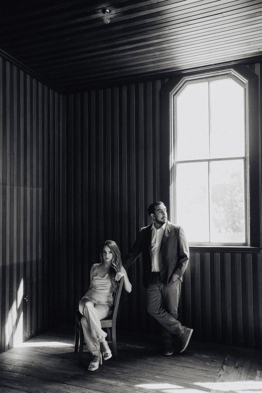 Couple by a tall window in a dark paneled room, hard light falling across the floor