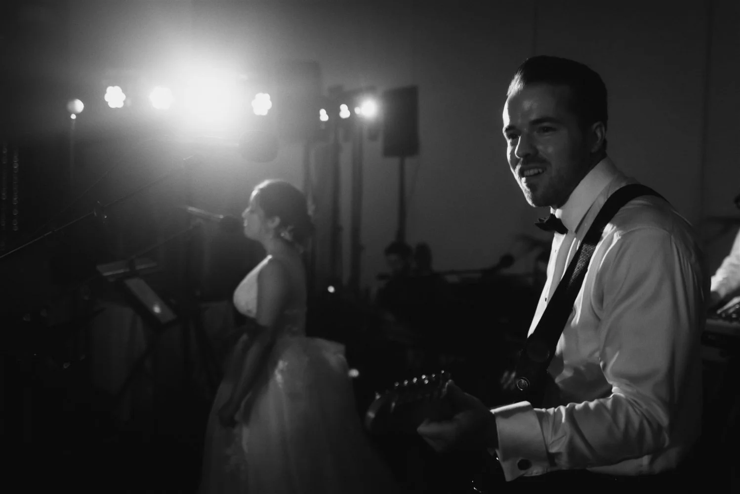 A groom in a tuxedo and bow tie playing electric guitar under stage lights at a wedding reception while the bride watches
