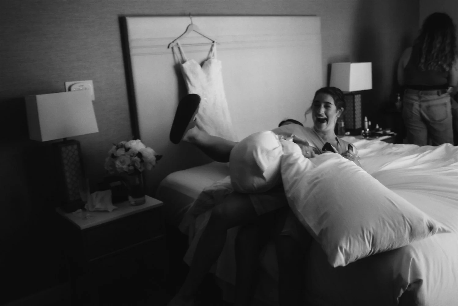 A bride lying back on a hotel bed laughing and holding a shoe, her wedding dress hanging on the headboard behind her