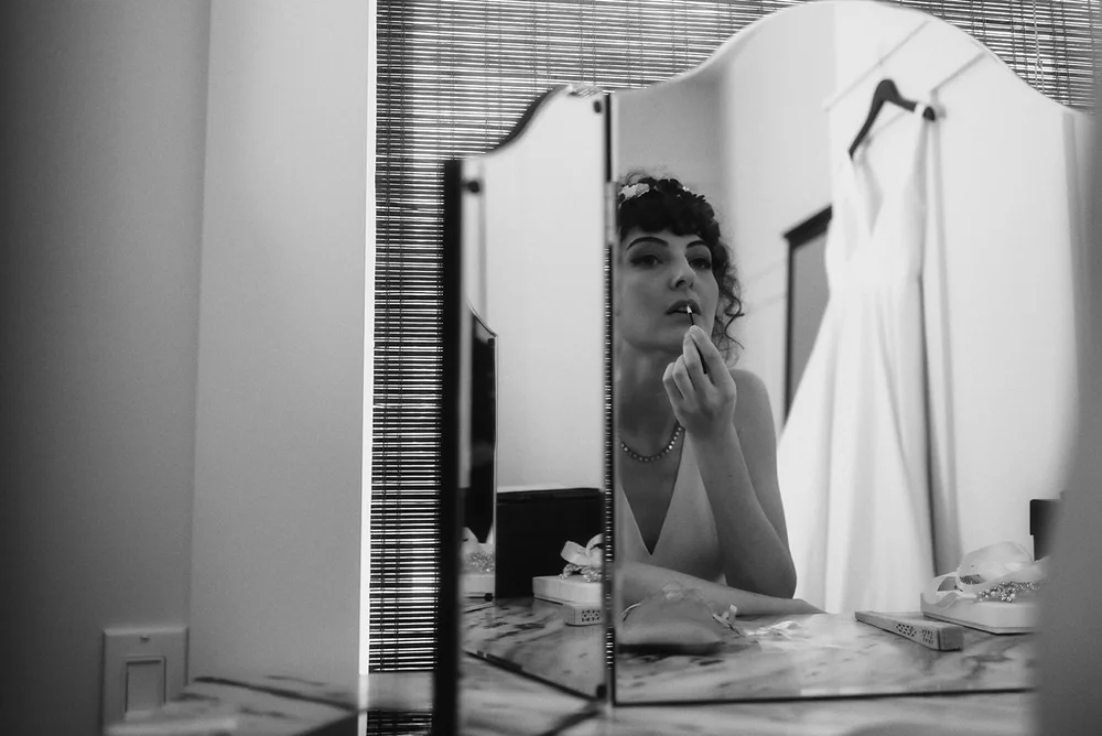 Bride putting on lipstick at a mirror, her gown hanging behind her