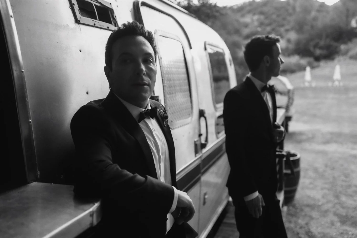 Black and white frame of a groom leaning against a silver vintage trailer bar
