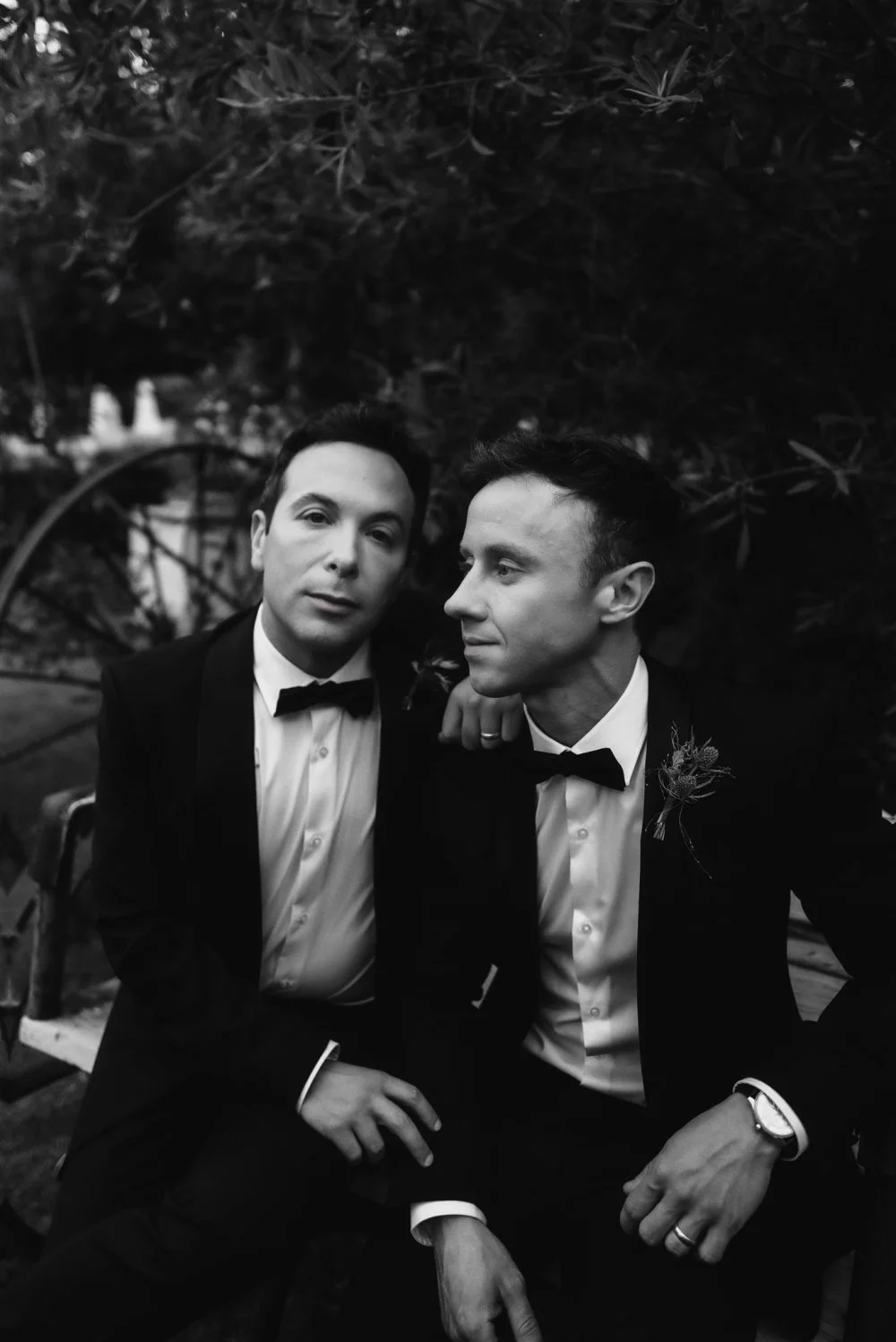 Two grooms in tuxedos sitting shoulder to shoulder outdoors, in black and white