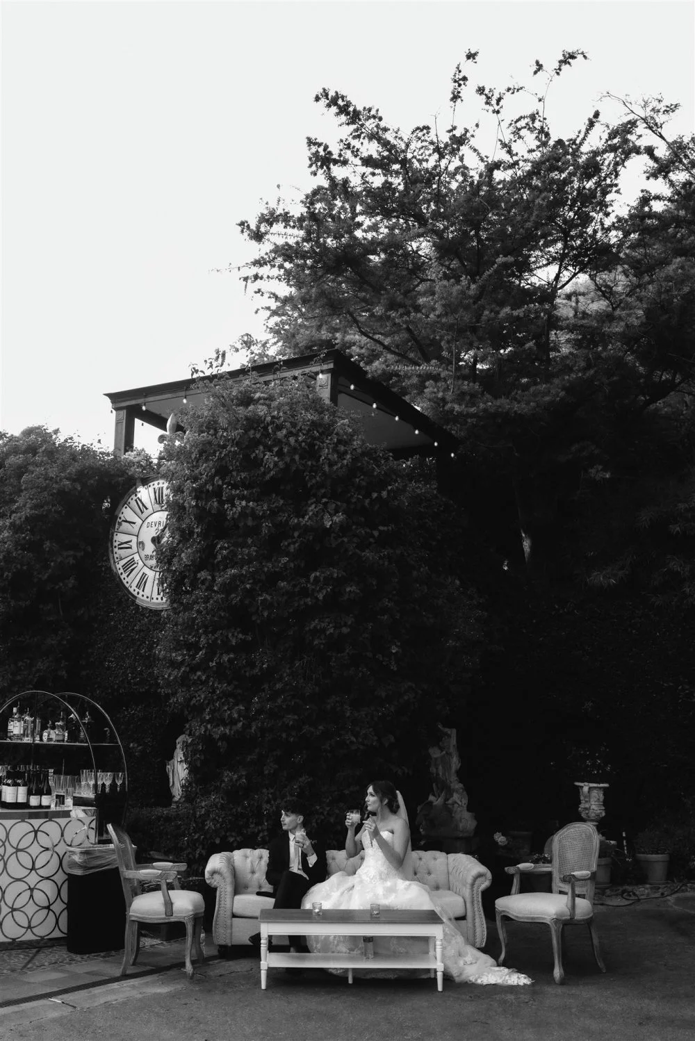 Bride and groom on a sofa in a garden lounge beneath old trees, in black and white