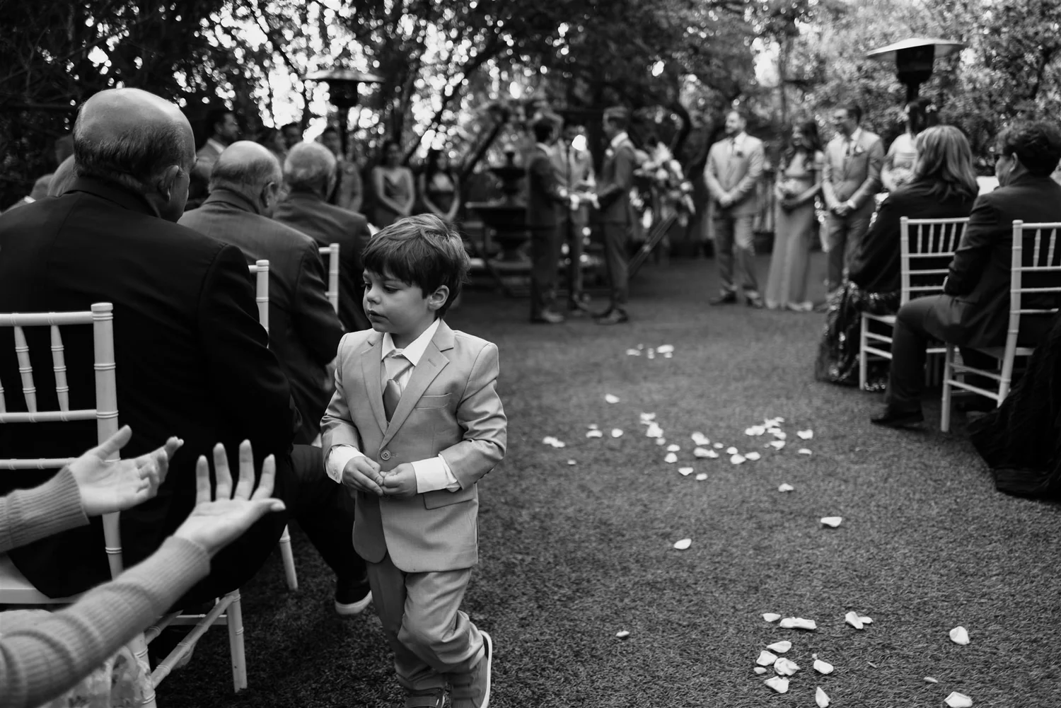 Black and white frame of a young ring bearer walking up a petal-strewn aisle between seated guests