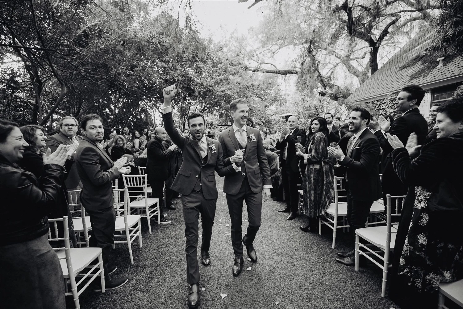 Two grooms walking back up the aisle at Green Gables Estate as their guests stand and cheer, in black and white