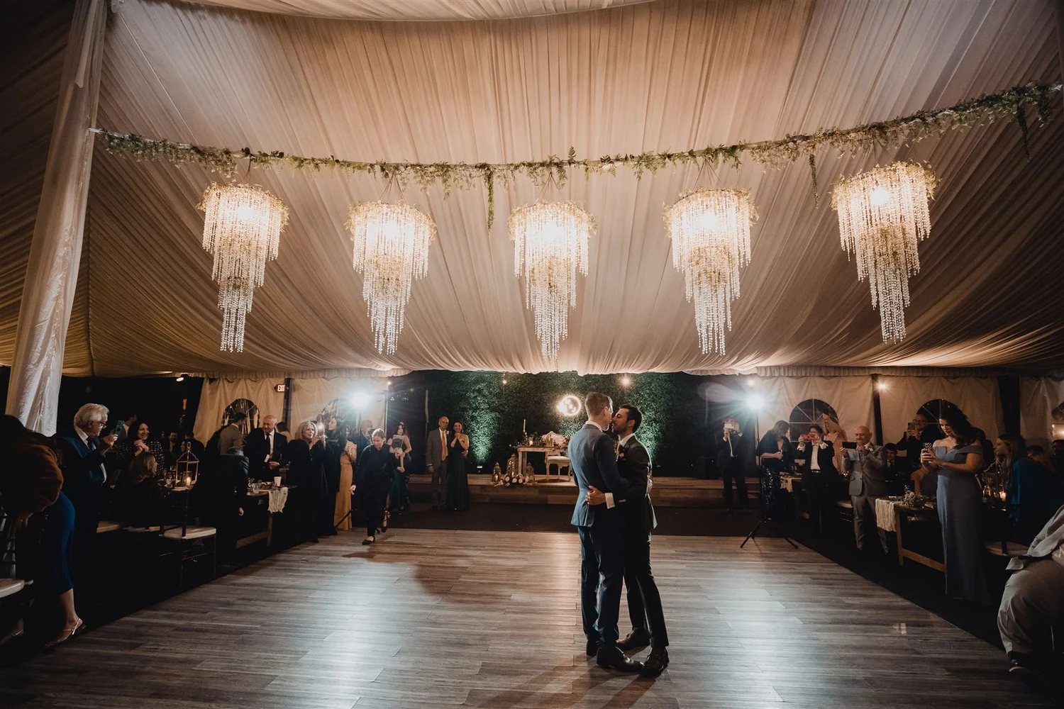 Two grooms sharing their first dance beneath crystal chandeliers in a draped reception tent