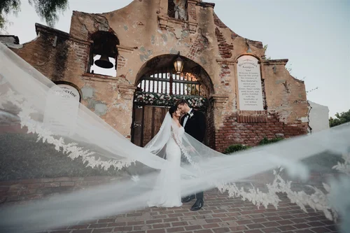 Bride and groom in an arched brick mission ruin, her embroidered veil sweeping across the frame