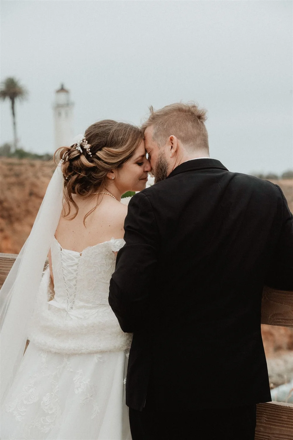 Bride and groom forehead to forehead, a lighthouse on the point behind them