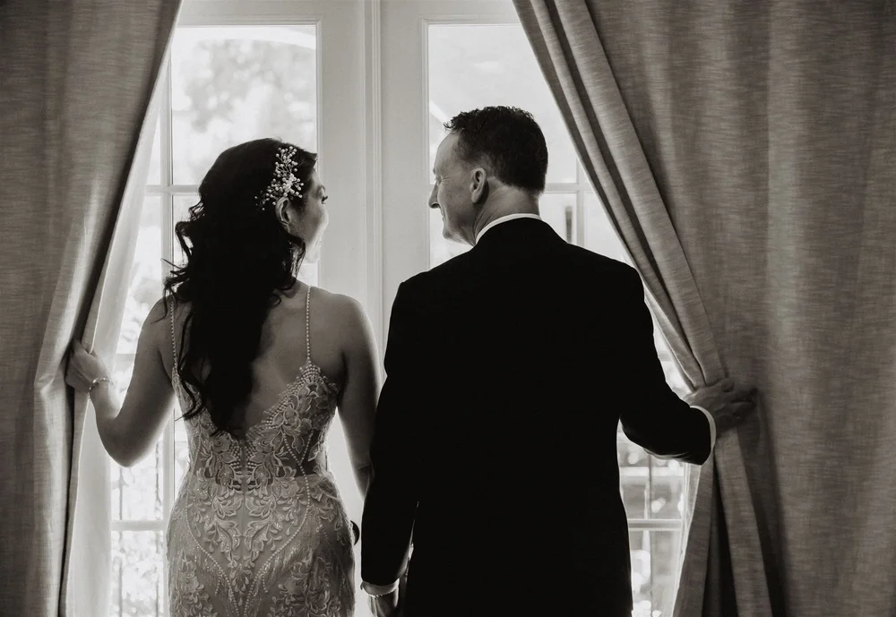 Bride and groom opening tall curtained windows, seen from behind, black and white
