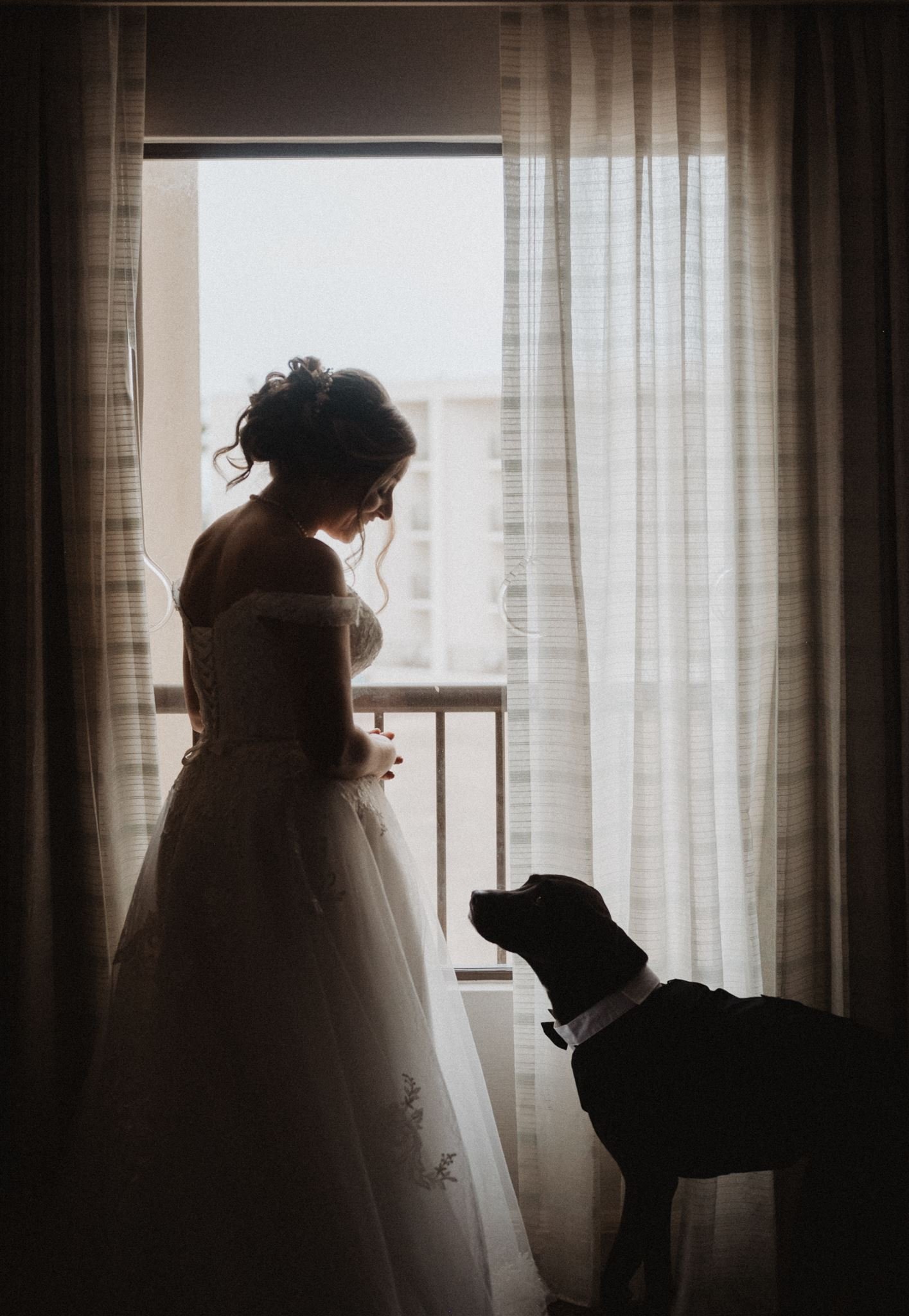 Bride standing by a curtained window with her dog beside her