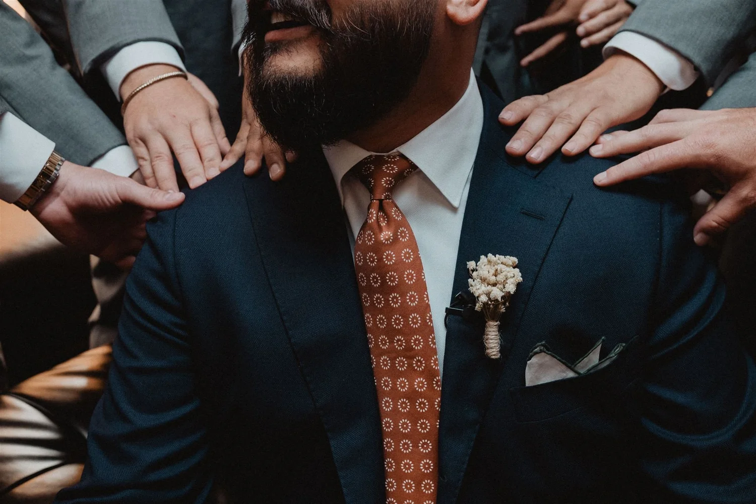 Groomsmen resting their hands on the groom’s shoulders, his orange patterned tie and boutonniere in the centre of the frame