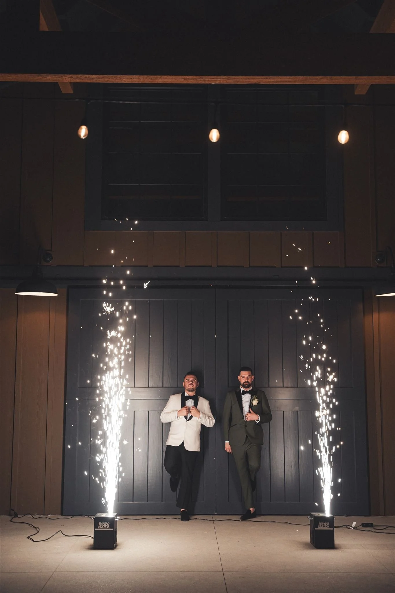 Two grooms walking out of a barn doorway between two cold-spark fountains at the end of the night