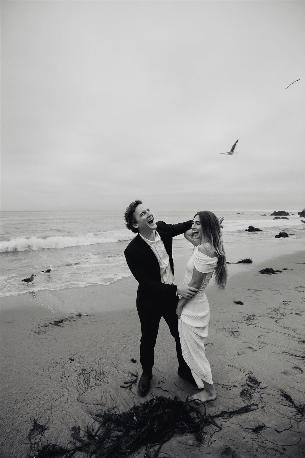Black and white photo of an engaged couple laughing together on the beach