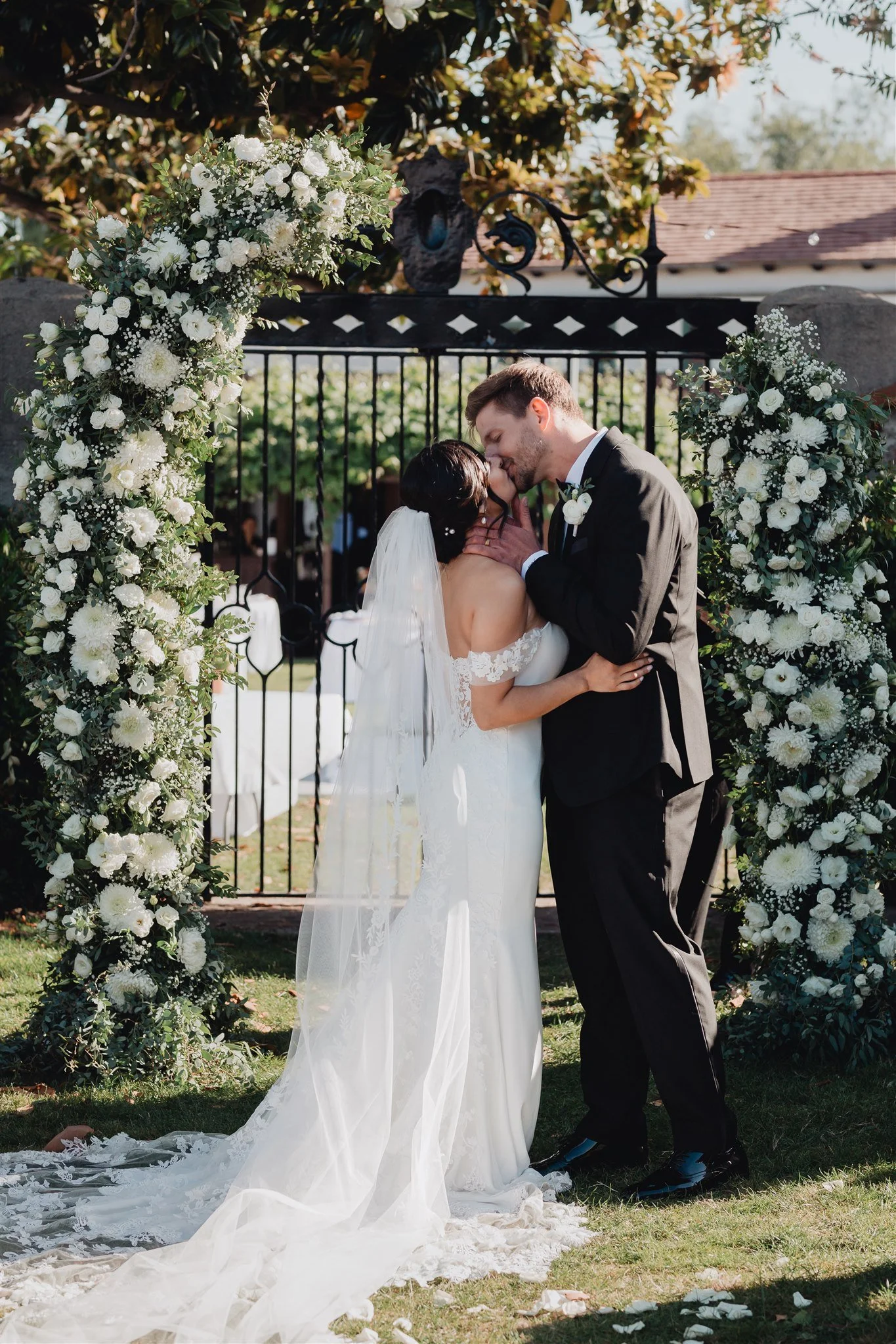 Couple kissing under a white floral arch at the end of the ceremony at Grapevine Arbor