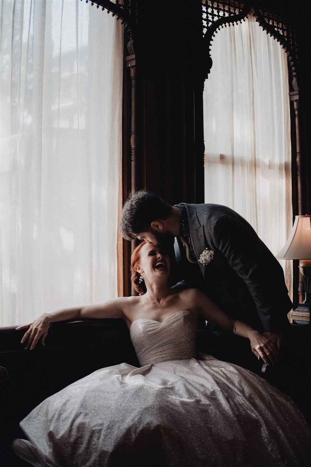 A bride laughing in her gown beside a curtained window while her groom leans over her