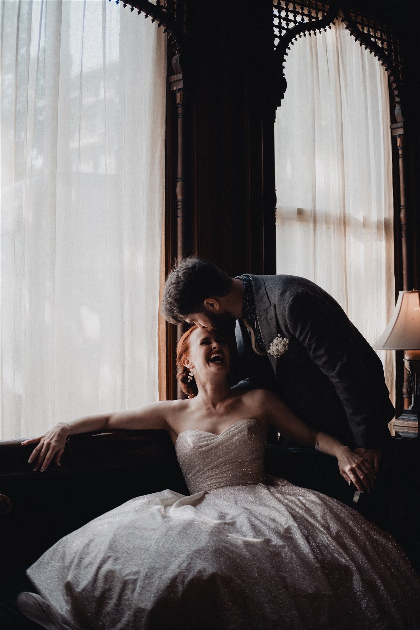 A laughing bride with red hair on a sofa in a window-lit room, her groom leaning over her, photographed at a Los Angeles wedding